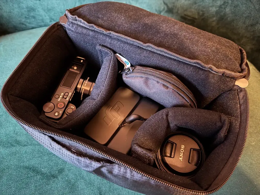 A picture of a camera, a lens, a case for batteries and a case for microphones in a soft gray pouch. The pouch is on a green sofa.