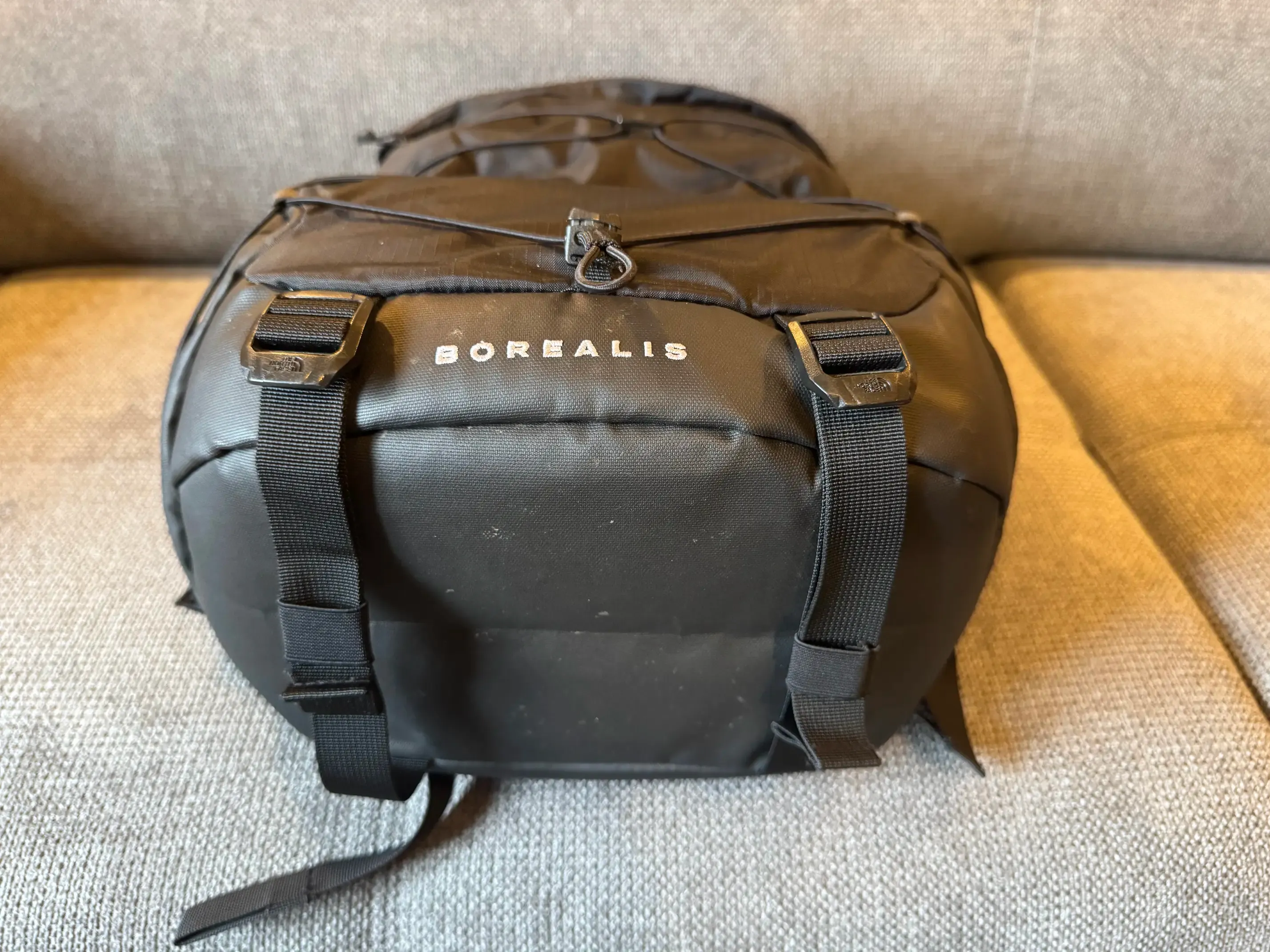 A photo of the bottom of a black backpack. We can see two straps that can be tightened to flatten the backpack.