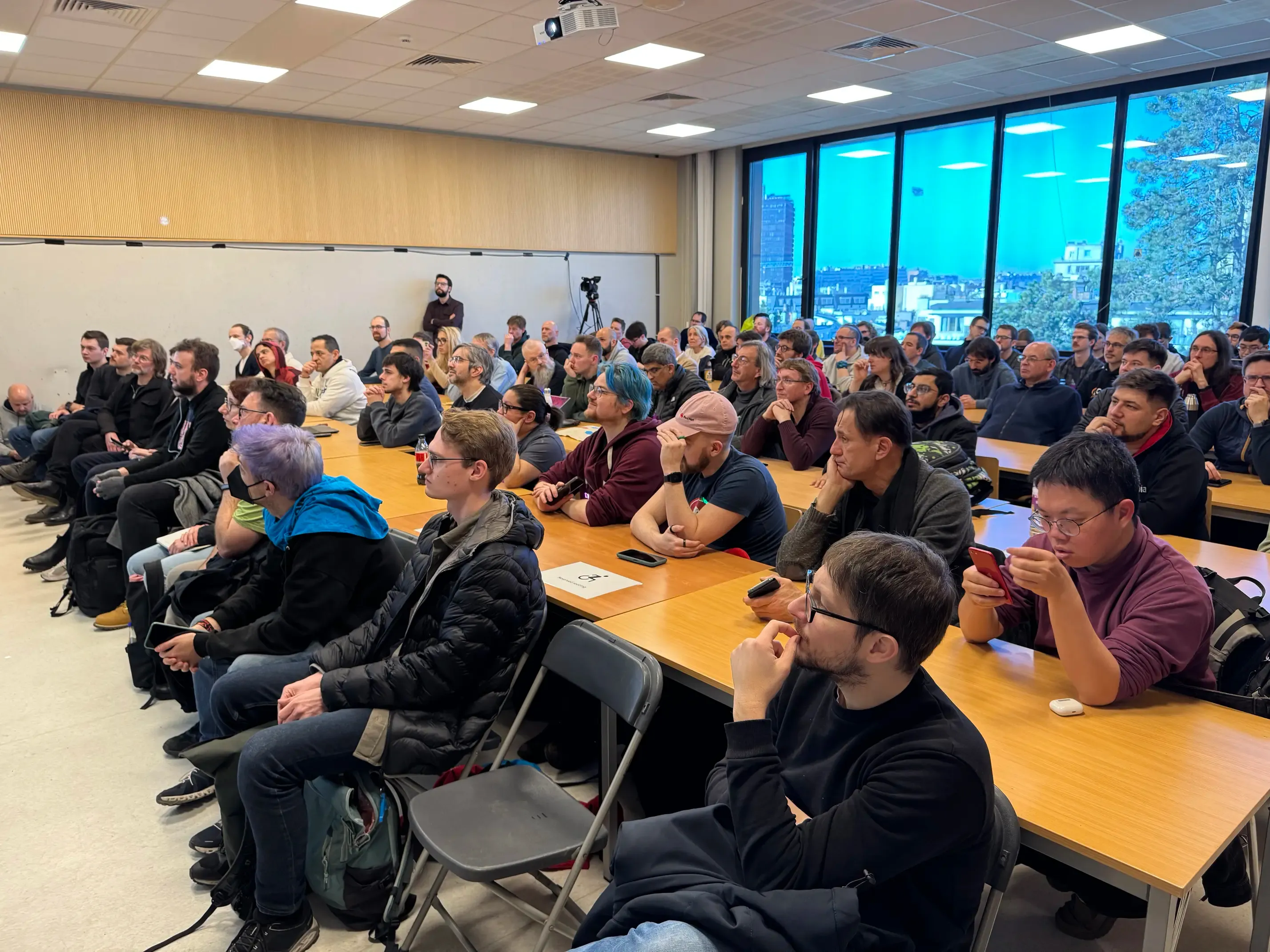 A photo of a university room full of people listening to a speaker that we can't see.