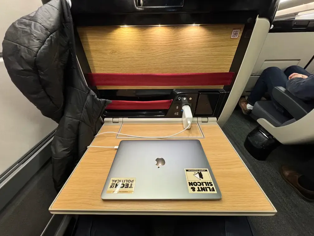 A photo taken from a train seat in first class. There is a large tablet on which is a MacBook Pro laptop. There is a charging block plugged to the seat.